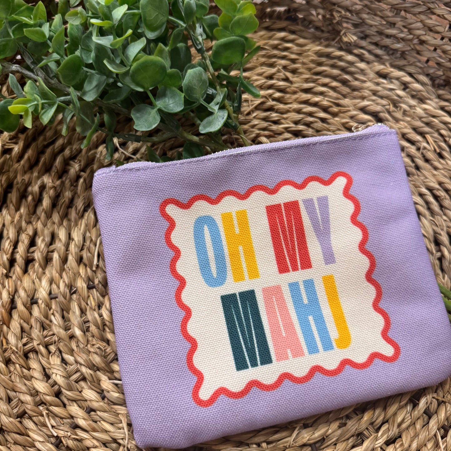 A lavender canvas zippered pouch with a retro-style graphic that reads 'OH MY MAHJ' in multicolored letters on a cream background with a red wavy border, resting on a woven straw mat next to a green plant.