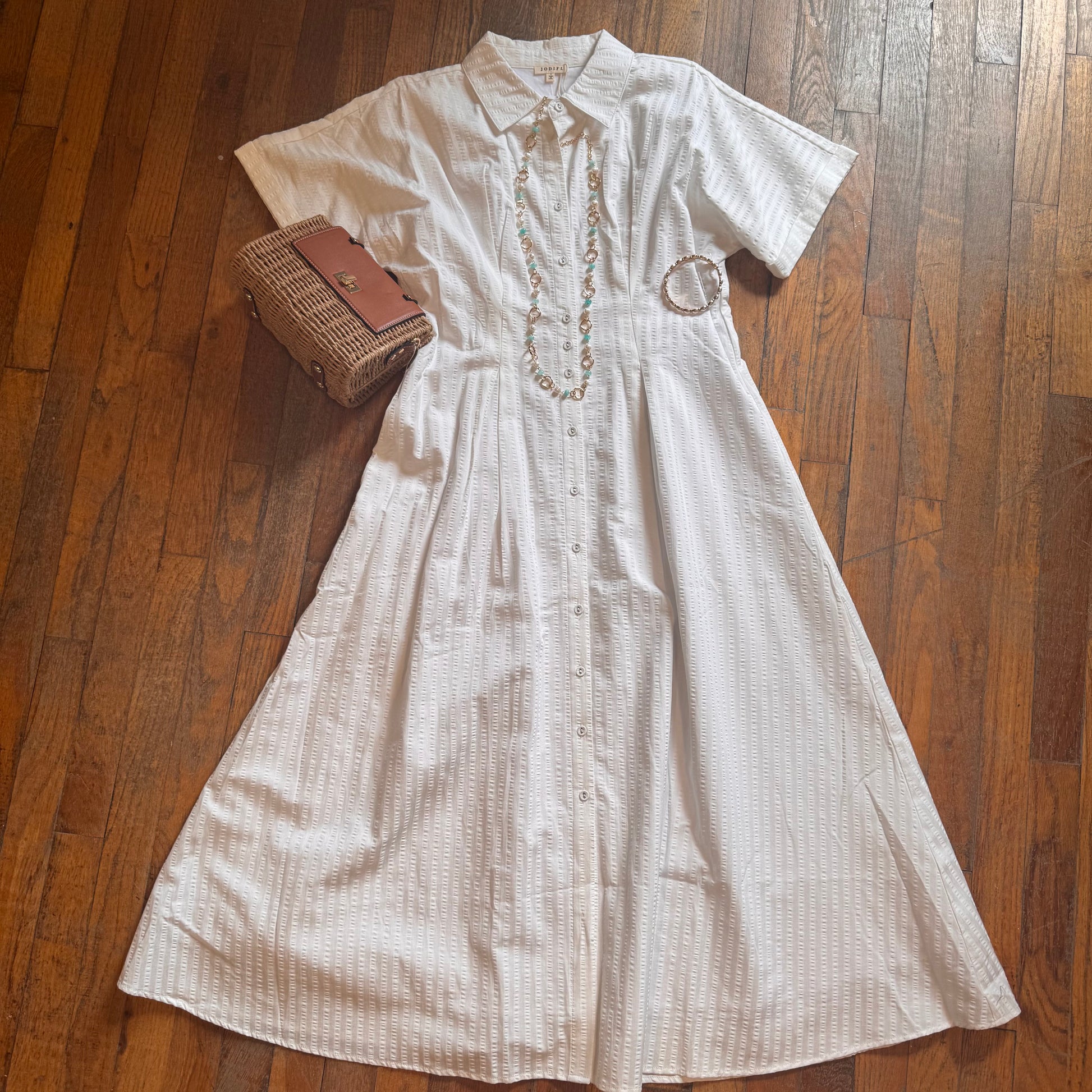 White dress with embroidery on a wooden floor, accompanied by a woven handbag and necklace.