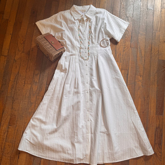 White dress with embroidery on a wooden floor, accompanied by a woven handbag and necklace.