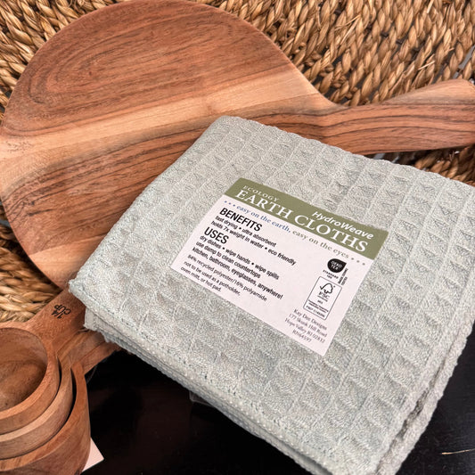 Wooden cutting board, wooden measuring spoons, and a gray cloth with a label on a woven mat.