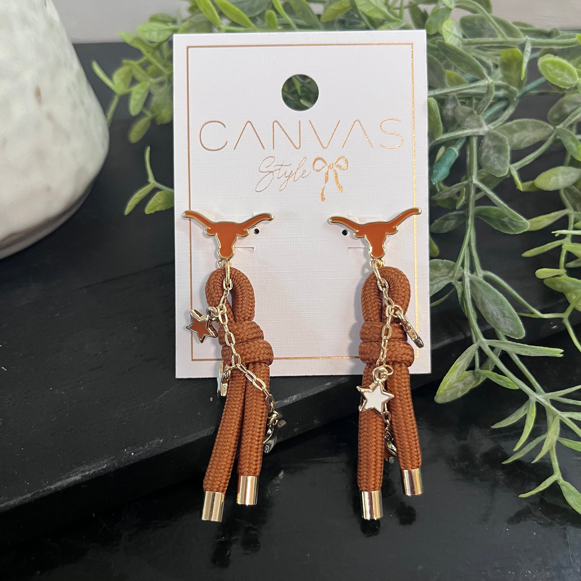 Brown earrings with gold accents on a 'Canvas Style' branded card, set against a dark surface with greenery.