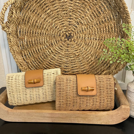 Two woven bags with brown leather accents on a wooden tray, with a large woven basket in the background.