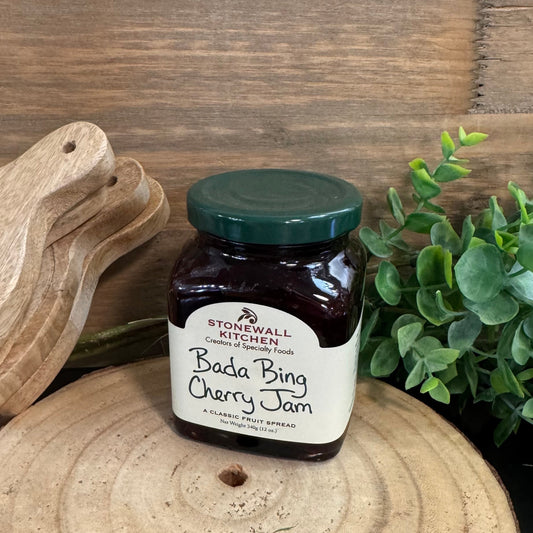 Jar of Stonewall Kitchen Bada Bing Cherry Jam on a wooden surface with a wooden cutting board and plant in the background.