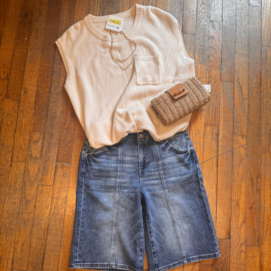 Beige sleeveless top, blue denim shorts, and woven clutch on a wooden floor