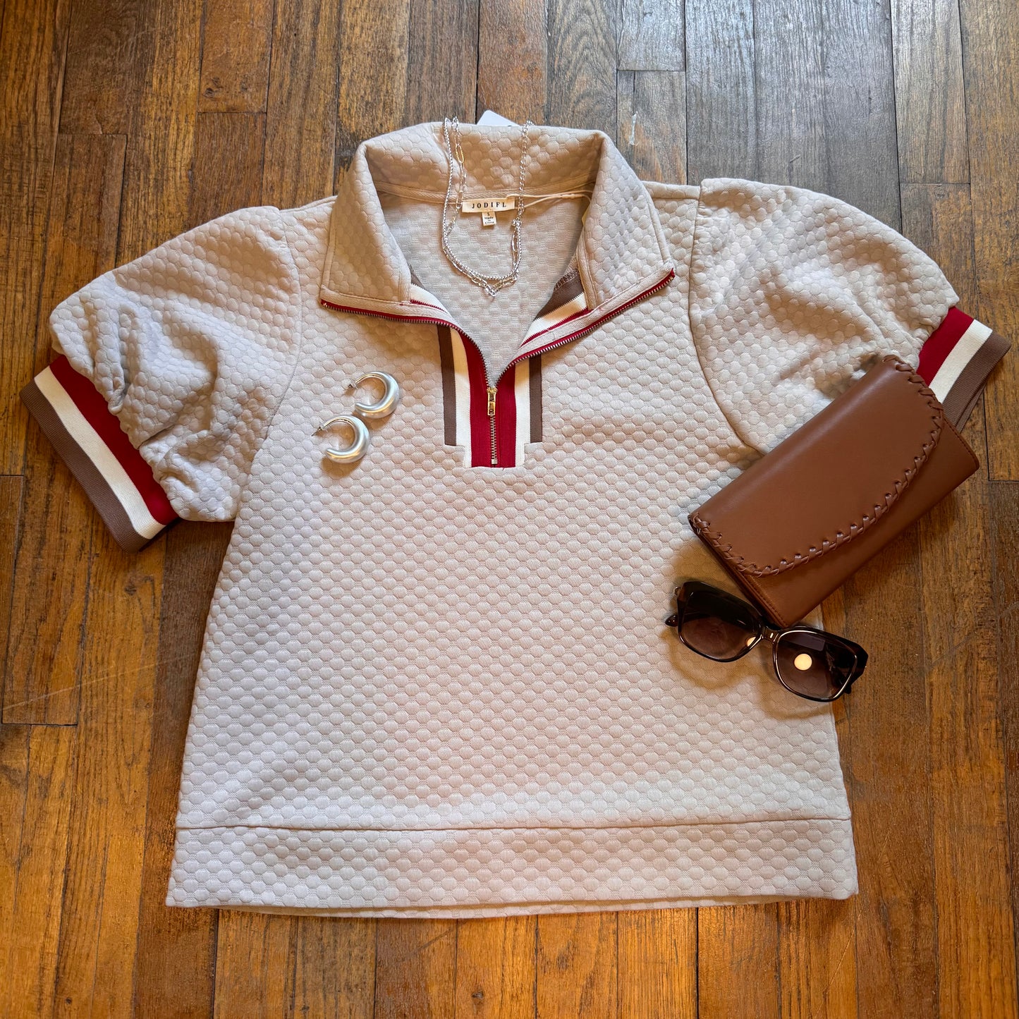 Light pink quilted jacket with red and white accents on a wooden surface, accompanied by a brown wallet and sunglasses.