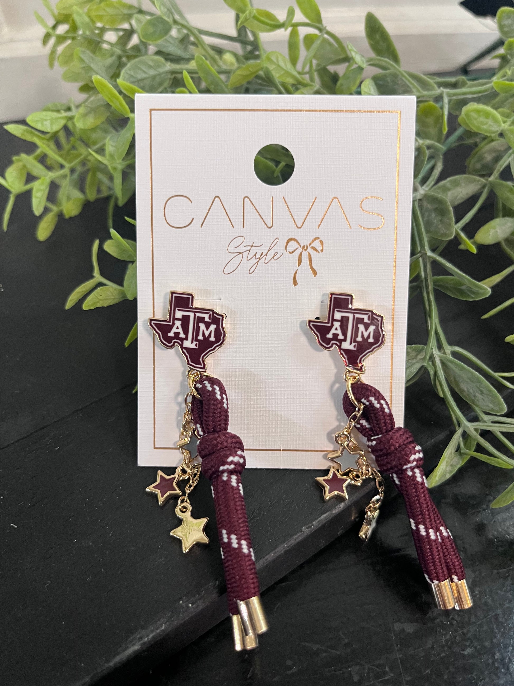 Maroon hair ties with gold charms on a 'Canvas Style' branded card, set against a dark surface with greenery.