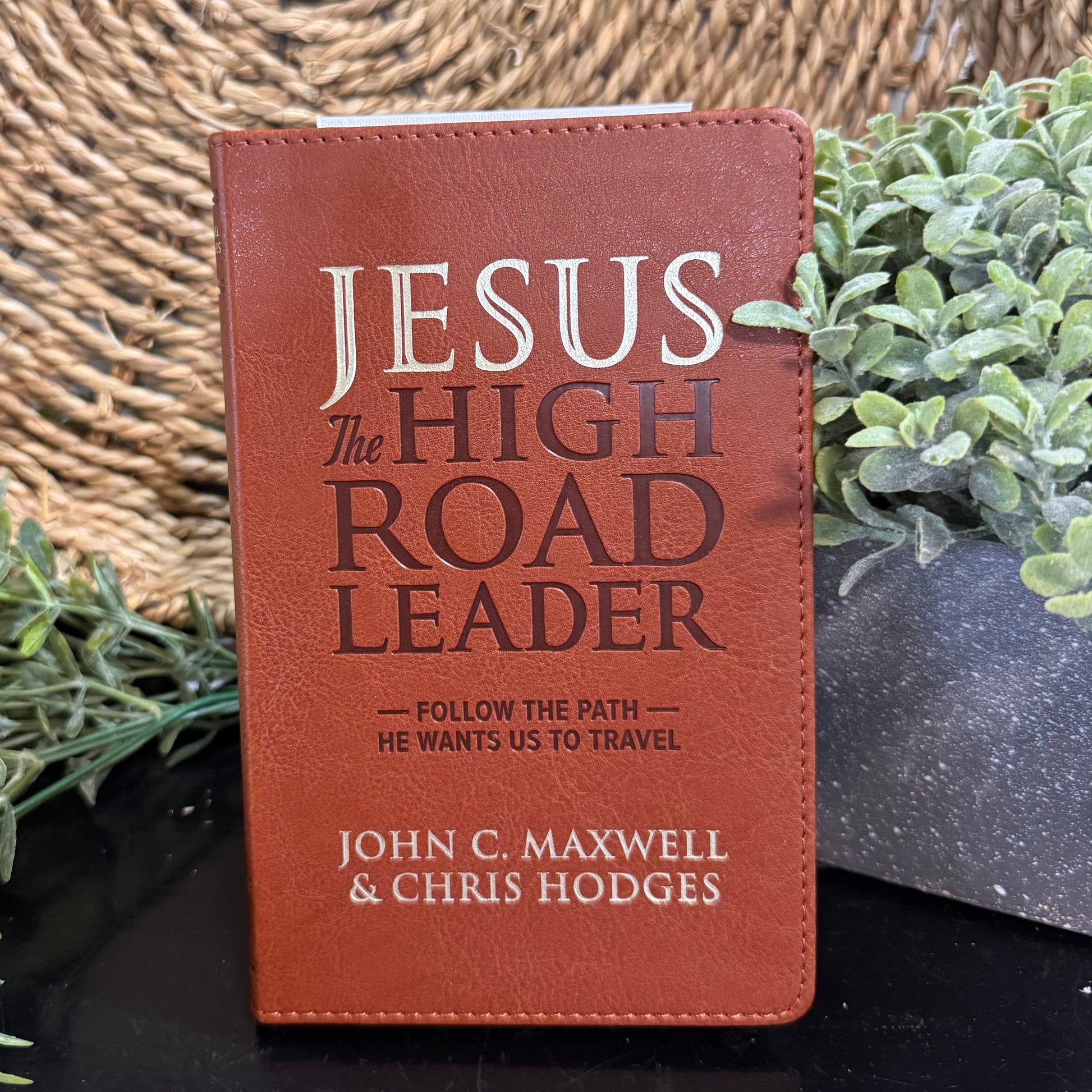 Brown book titled 'Jesus The High Road Leader' by John C. Maxwell & Chris Hodges, surrounded by plants.