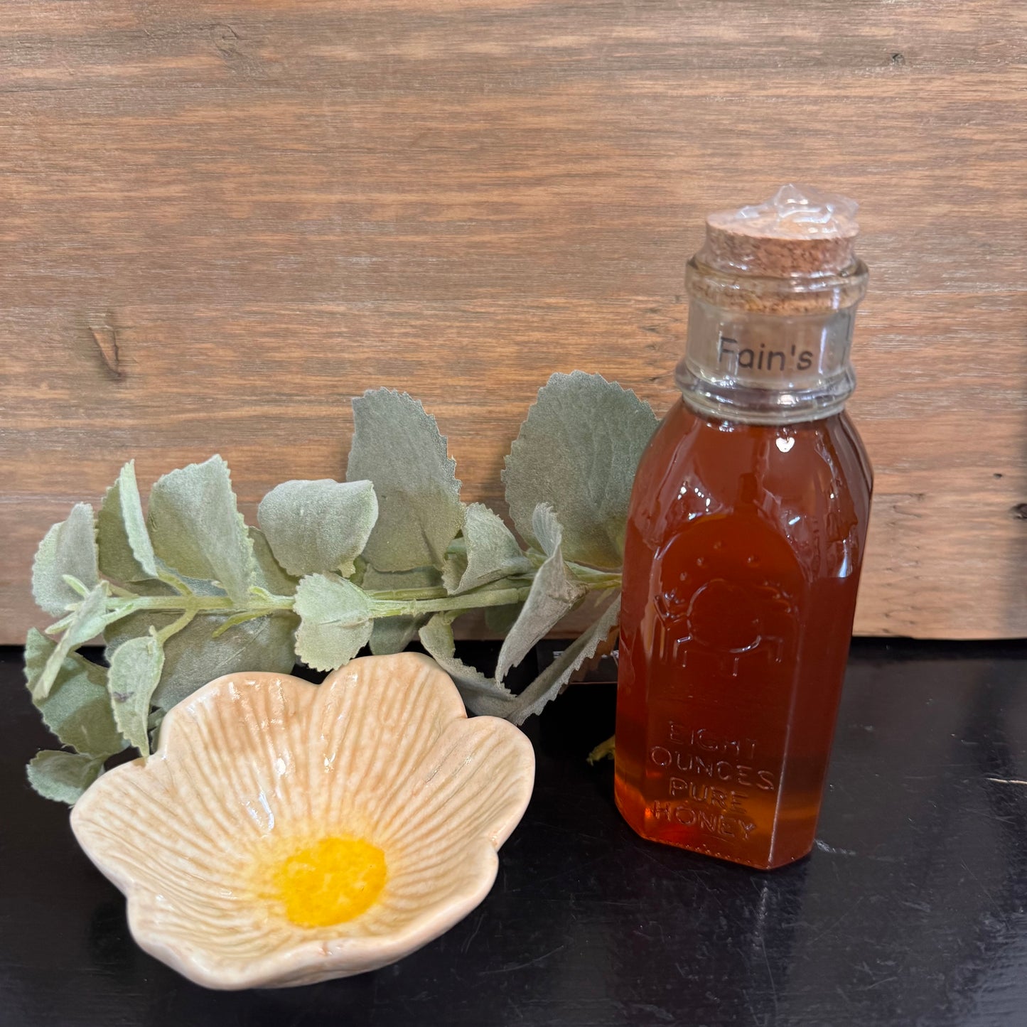 A small glass bottle filled with amber honey sits on a dark surface beside a flower-shaped dish with yellow wax and green leaves, set against a rustic wood background.
