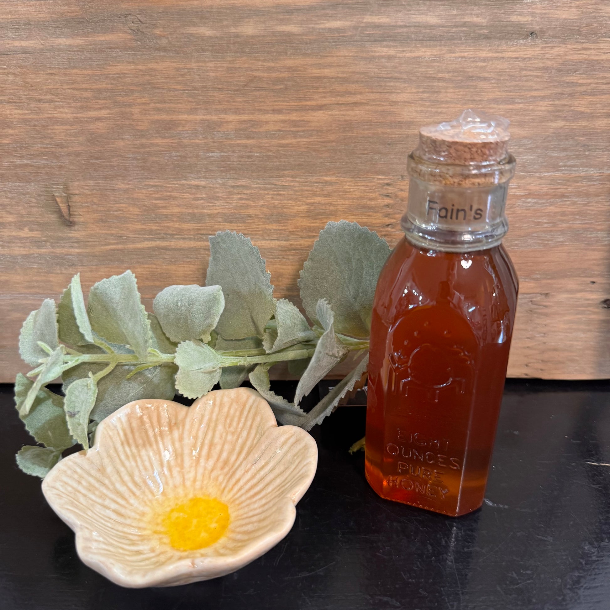 A small glass bottle filled with amber honey sits on a dark surface beside a flower-shaped dish with yellow wax and green leaves, set against a rustic wood background.