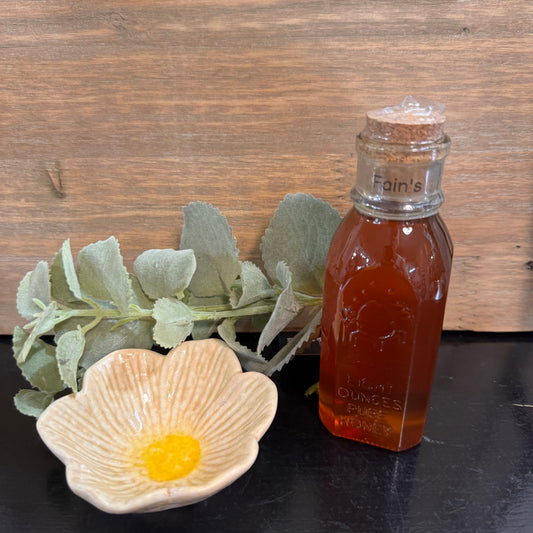 A small glass bottle filled with amber honey sits on a dark surface beside a flower-shaped dish with yellow wax and green leaves, set against a rustic wood background.