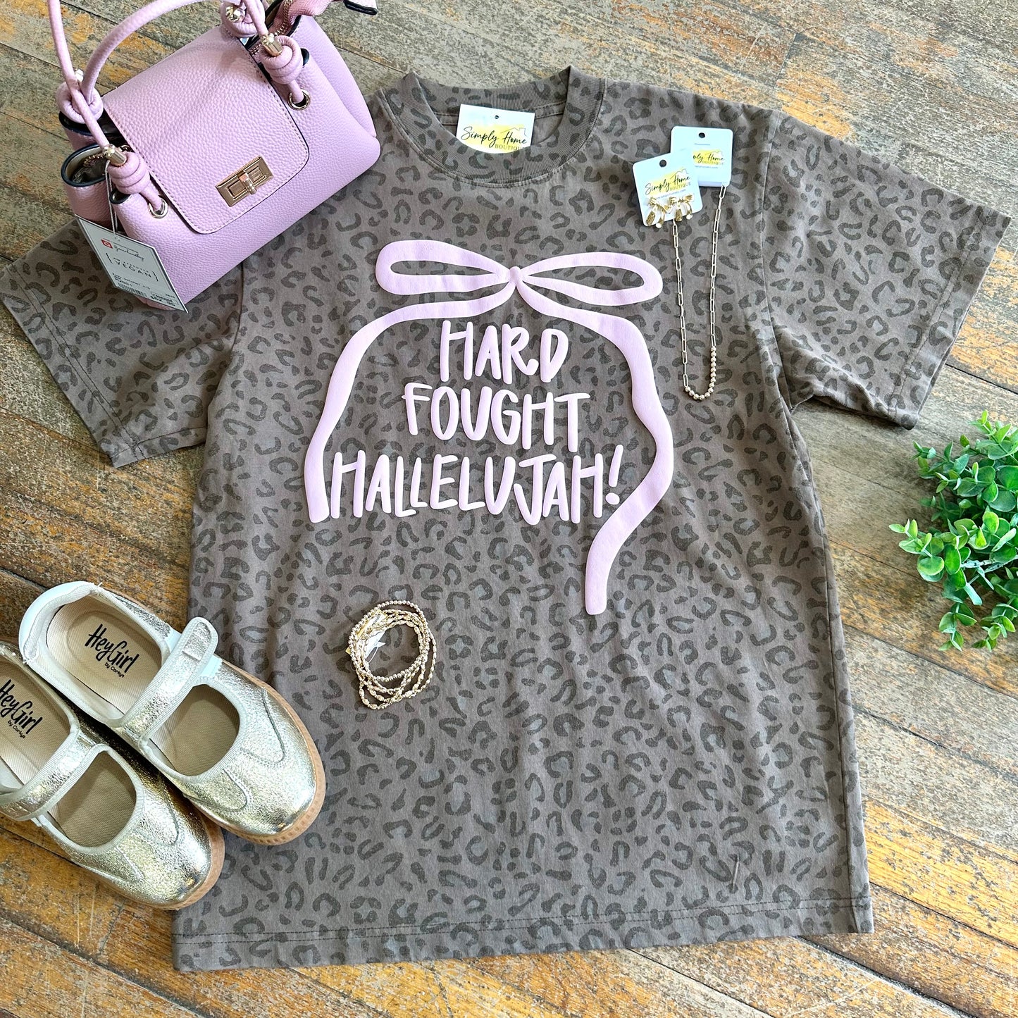 Gray leopard print t-shirt with 'Hard Fought Hallelujah!' text, paired with shoes and a handbag on a wooden surface.