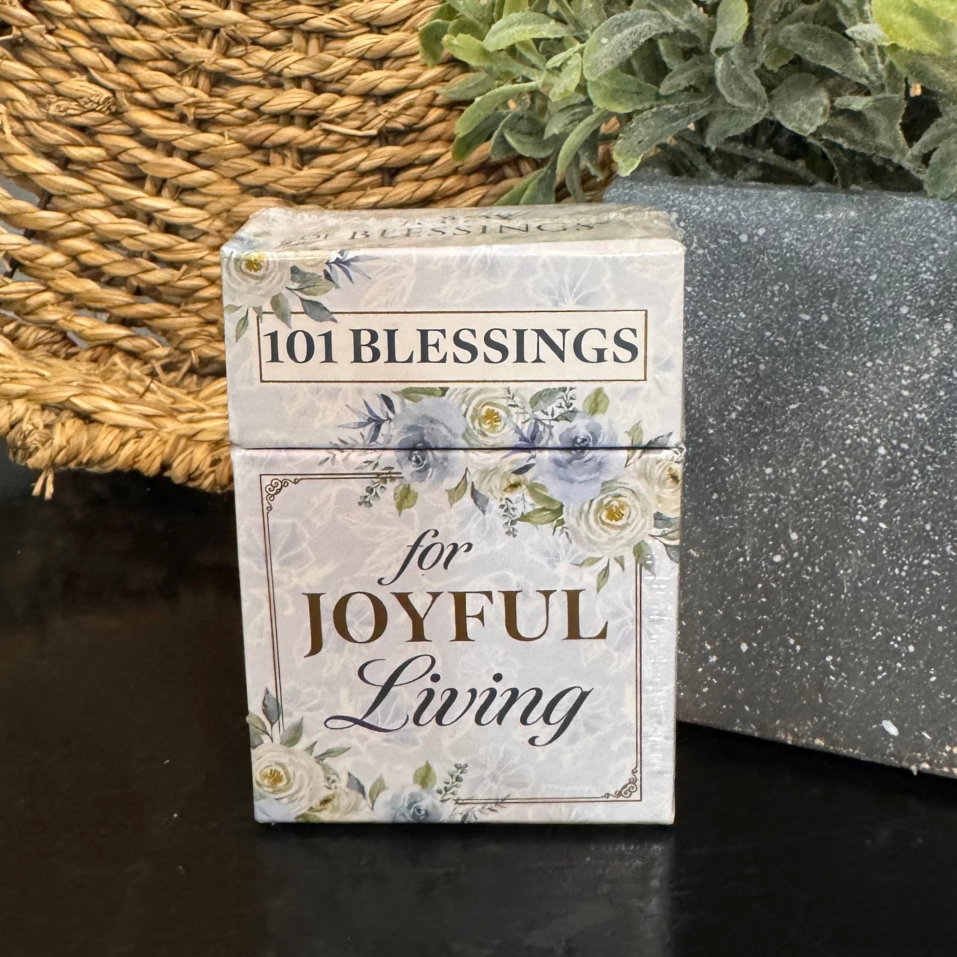 Small book titled '101 Blessings for Joyful Living' with floral design on a dark surface next to a woven mat and plant.