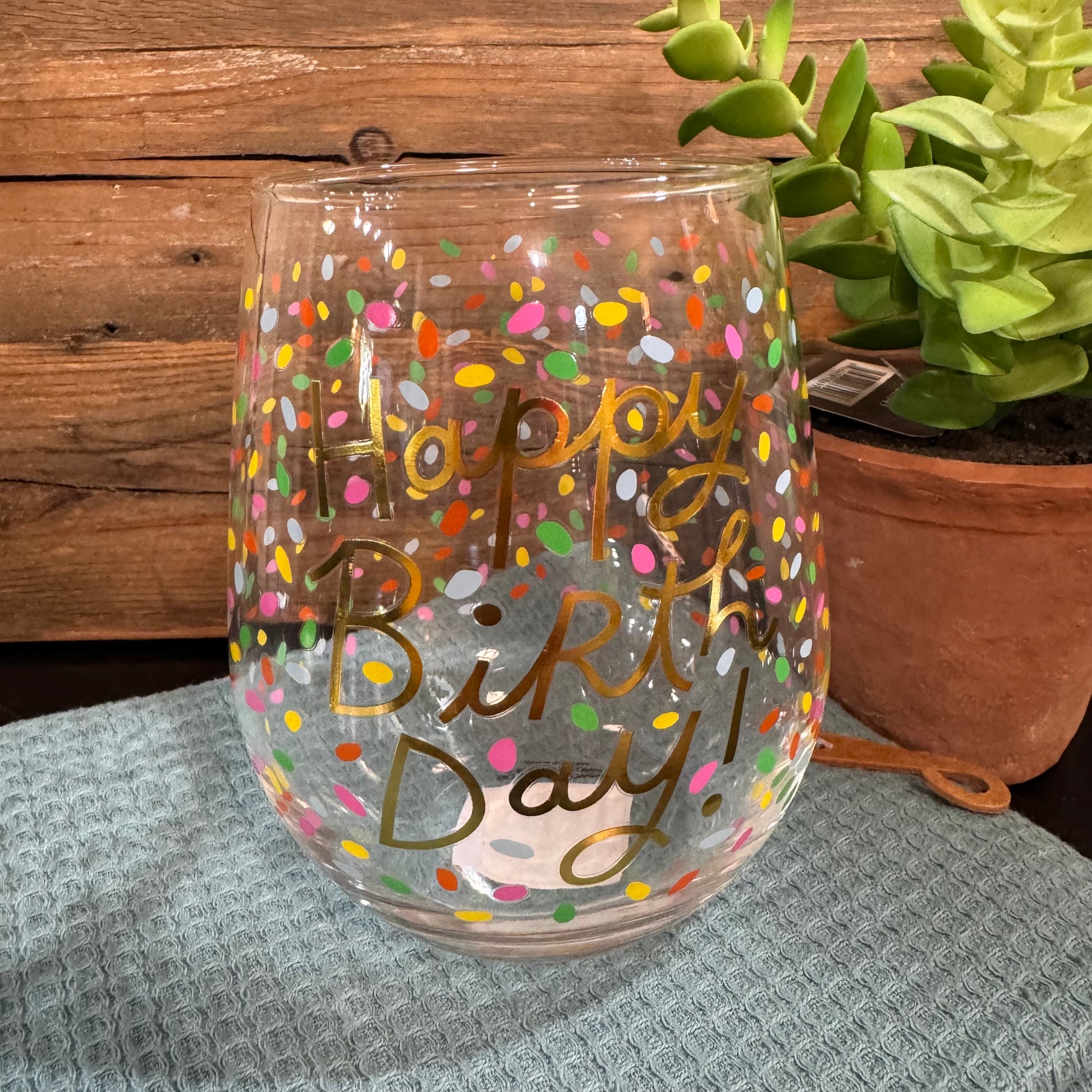 A clear stemless wine glass with gold foil lettering that reads 'Happy Birthday!' and decorated with colorful confetti dots, set on a gray textured cloth with a wooden wall and a potted succulent in the background.
