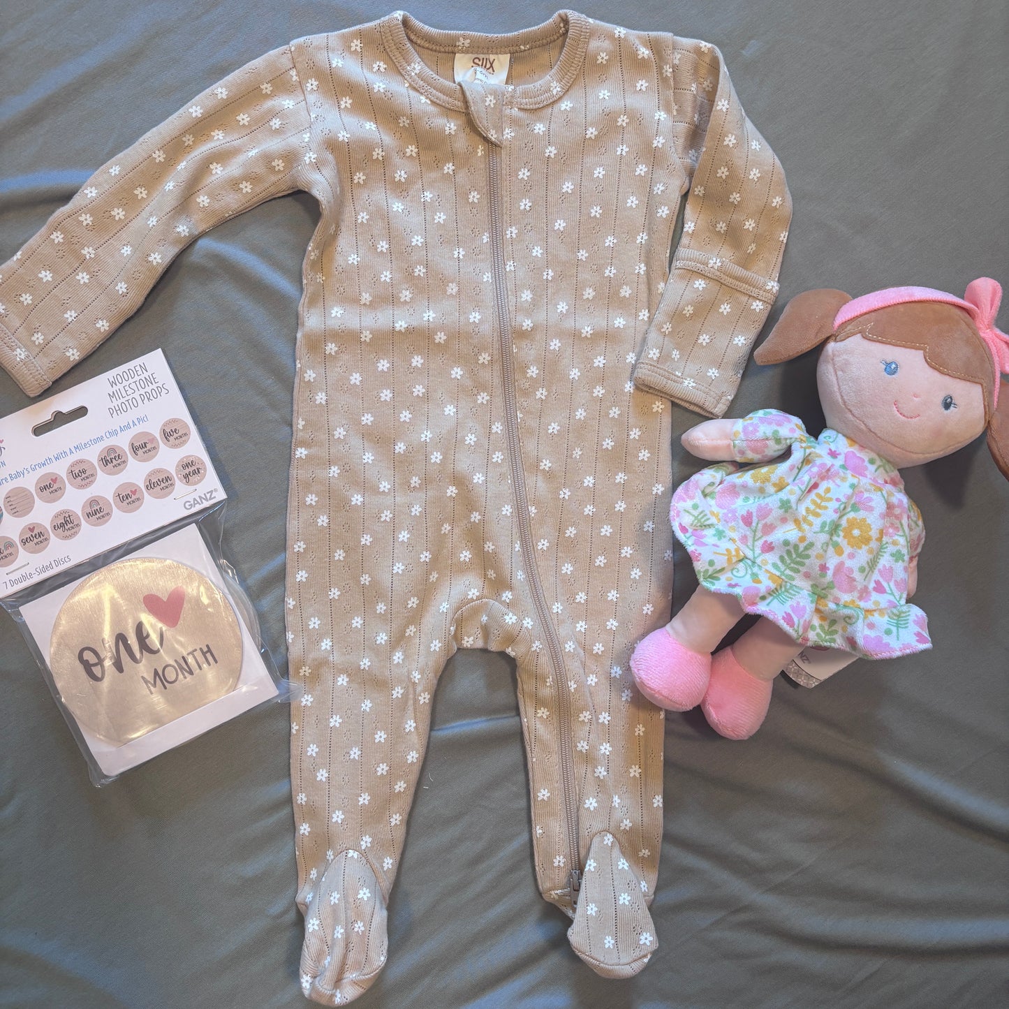 A baby footed sleeper in beige with white daisy patterns, laid flat on a gray fabric background next to a 'One Month' hat in its packaging and a plush doll.