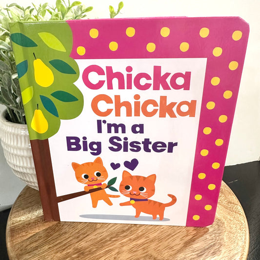 Children's book titled 'Chicka Chicka I'm a Big Sister' on a wooden surface with a plant in the background.