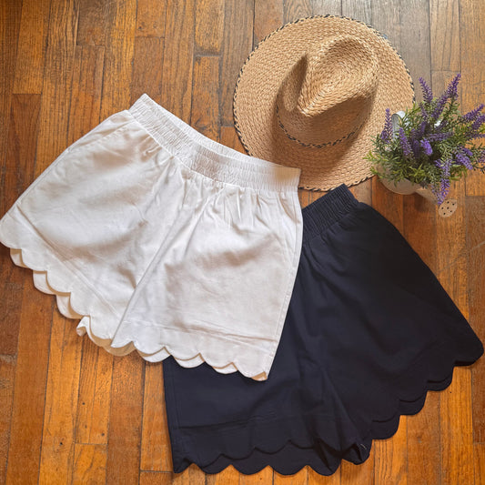 Two pairs of shorts and a straw hat on a wooden surface with a small plant.