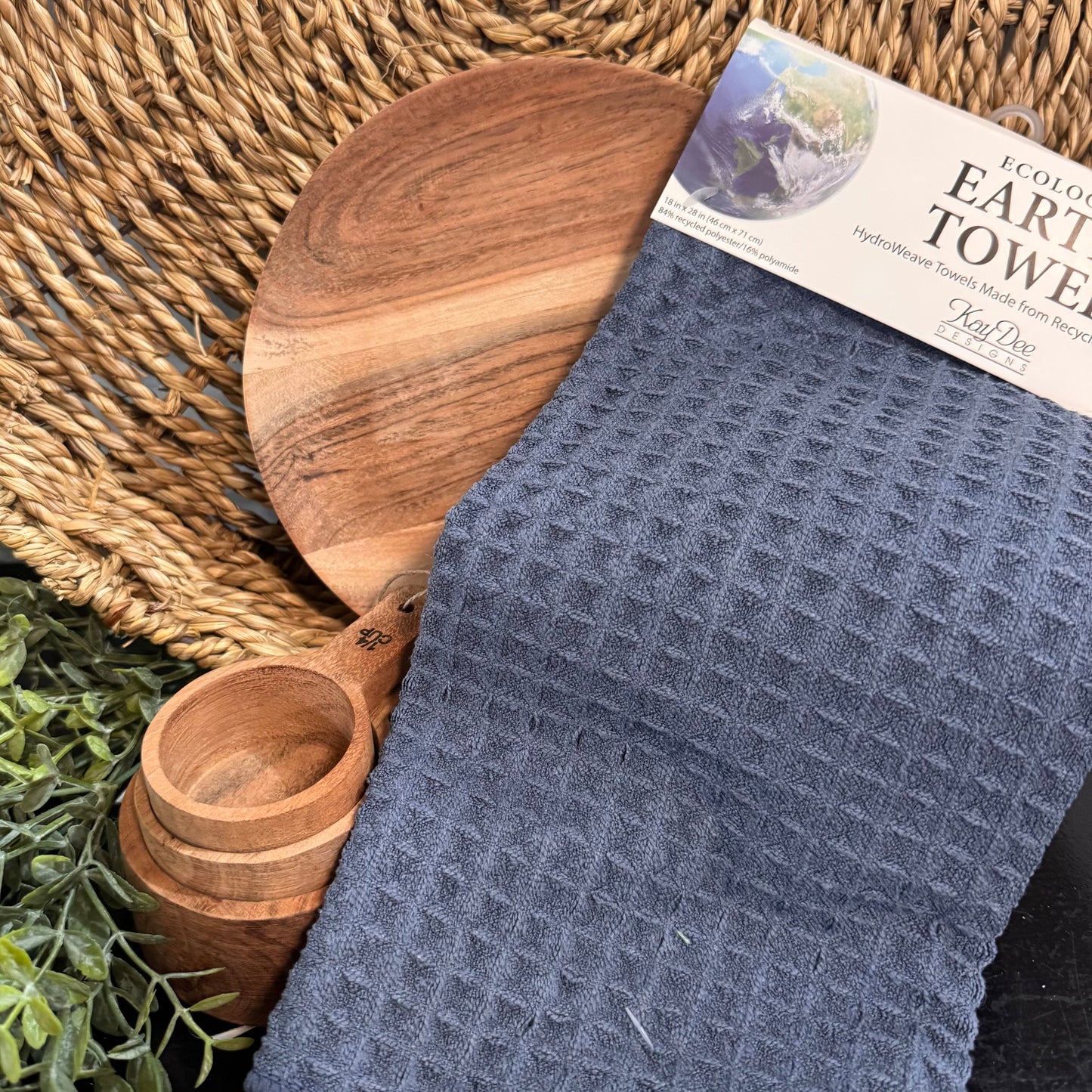 Blue towel with a label, wooden cutting board, and measuring cup on a woven mat with plants.