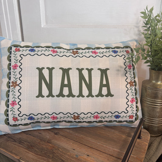 A rectangular decorative pillow with the word “NANA” in large dark green letters, surrounded by a floral embroidered border, resting on a rustic wooden bench in front of a white door with a potted plant nearby.