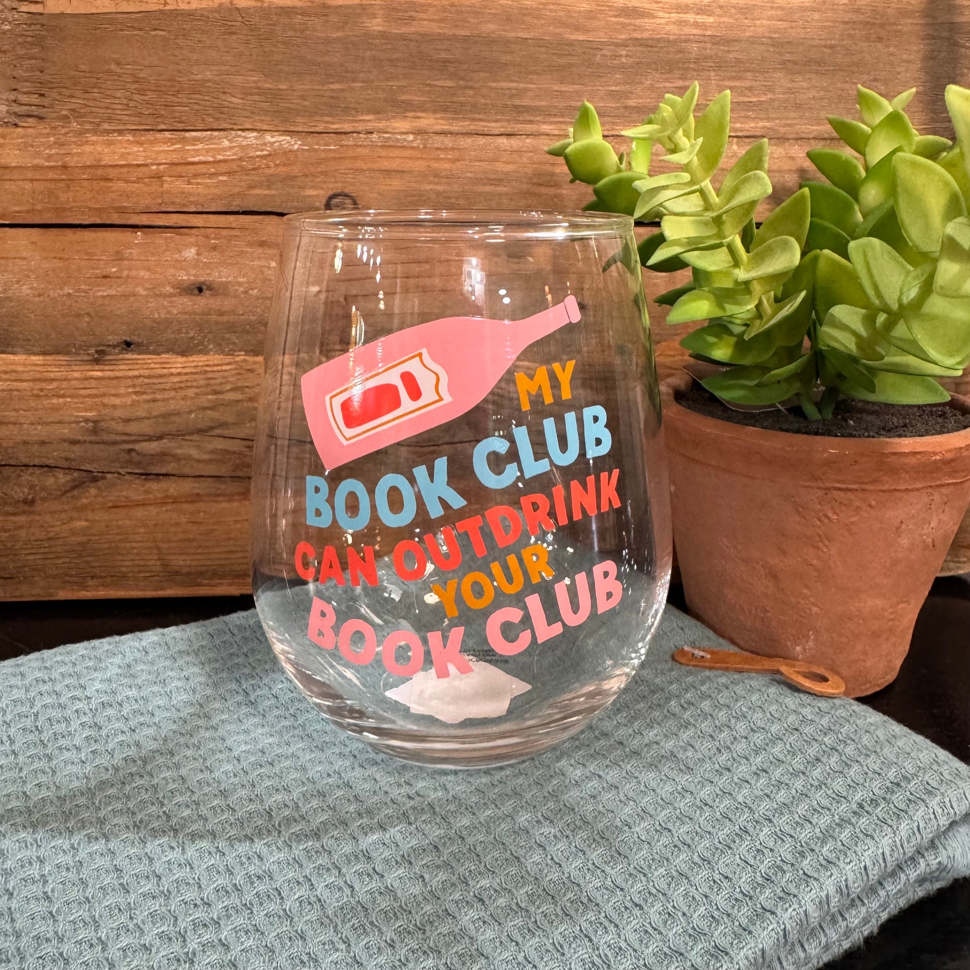 A clear stemless wine glass with colorful printed text reading 'MY BOOK CLUB CAN OUTDRINK YOUR BOOK CLUB' and a pink book illustration, placed on a gray textured cloth in front of a wooden wall with a potted succulent nearby.