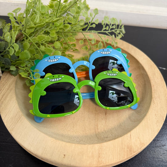 Children's sunglasses with dinosaur designs on a wooden surface with plants in the background