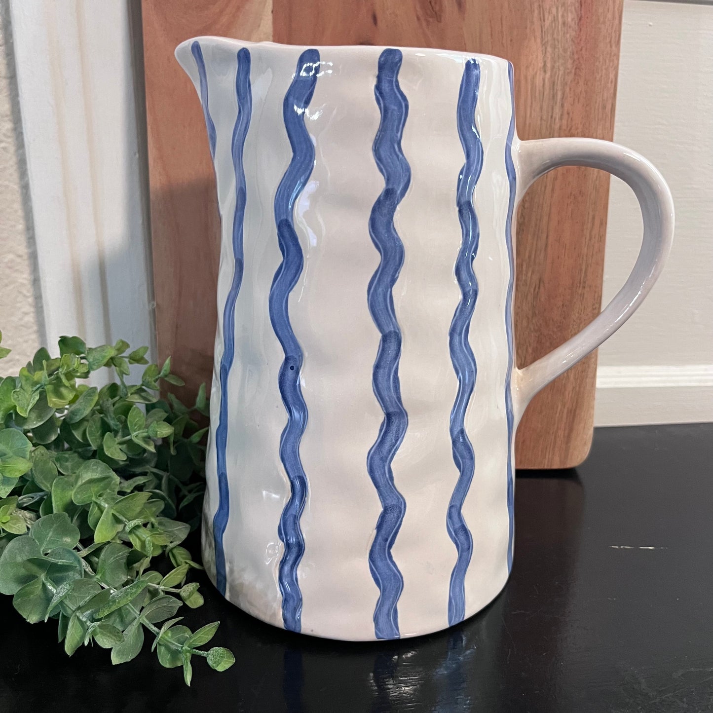 White ceramic pitcher with blue wavy patterns on a dark surface, next to a plant.