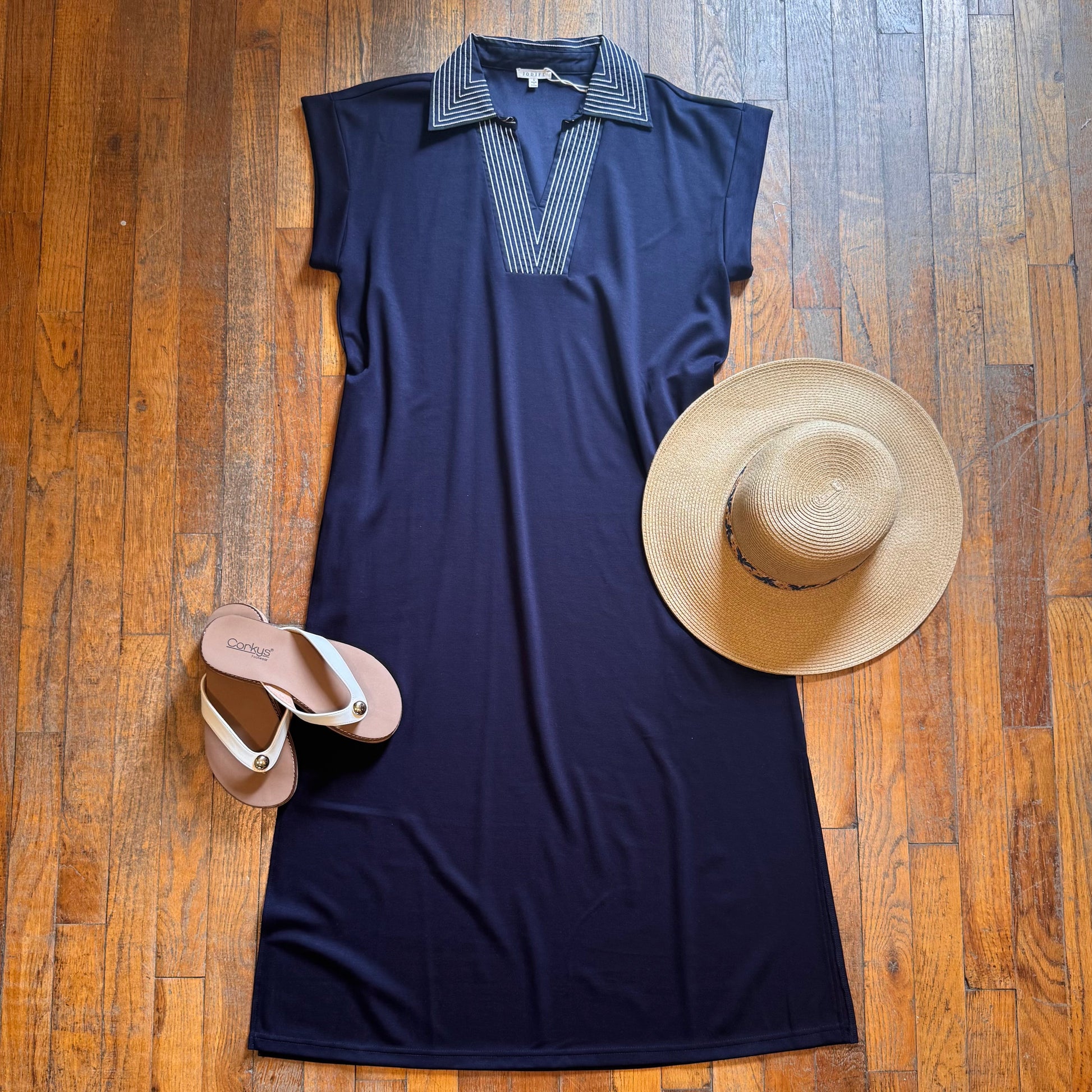 Navy sleeveless dress with striped collar, beige sandals, and straw hat on a wooden floor.