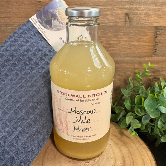 Bottle of Stonewall Kitchen Moscow Mule Mixer on a wooden surface with a towel and plant in the background.