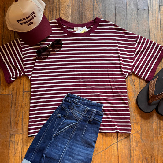 Maroon and white striped shirt with blue jeans, cap, sunglasses, and sandals on a wooden floor.