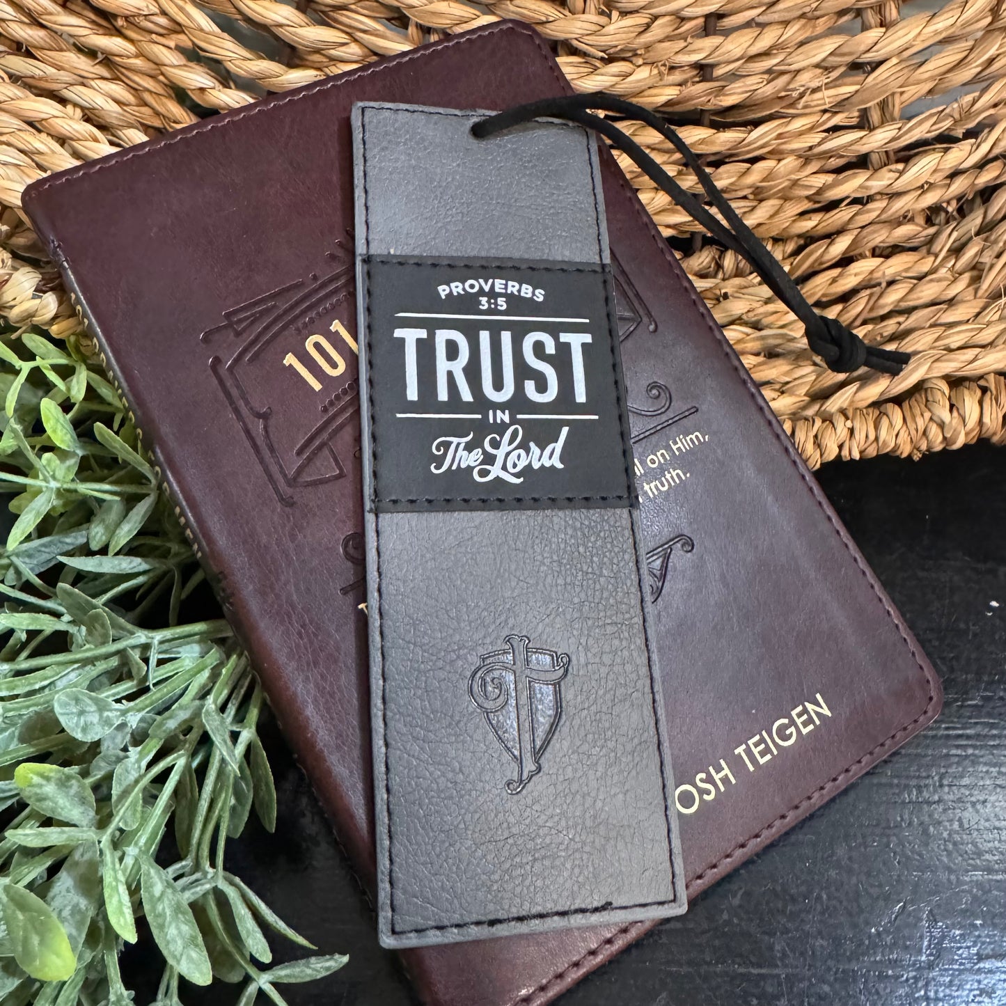 Brown leather book with a black and gray bookmark featuring 'Trust in the Lord' text, on a woven mat background.