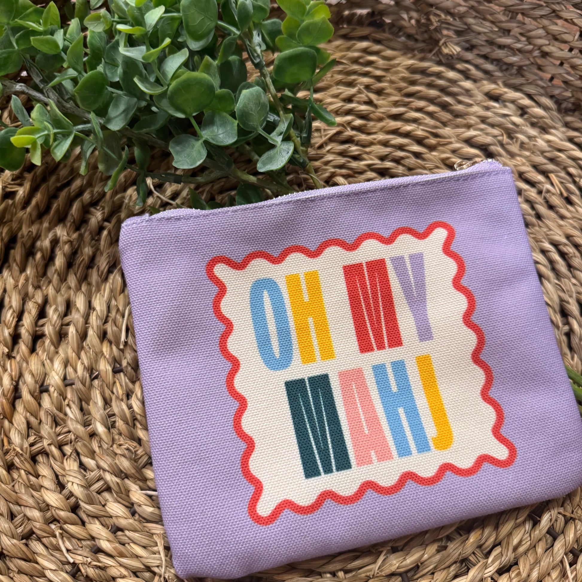 A lavender canvas zippered pouch with a retro-style graphic that reads 'OH MY MAHJ' in multicolored letters on a cream background with a red wavy border, resting on a woven straw mat next to a green plant.