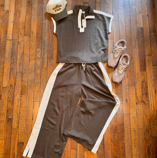 A flat-lay outfit arrangement on a wooden floor featuring a dark brown short-sleeve collared top with white trim, matching dark brown pants with white side panels, a beige and dark cap with 'coffee shop' text, and beige sneakers.
