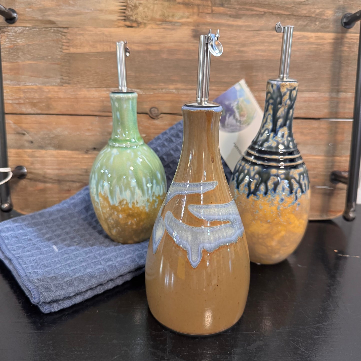 Three glazed ceramic oil bottles with metal pourers arranged on a dark tabletop against a rustic wood plank background. The front bottle is tan with a white and gray abstract design, the left bottle is light green with a gold base, and the right bottle is black and blue with a yellow base.