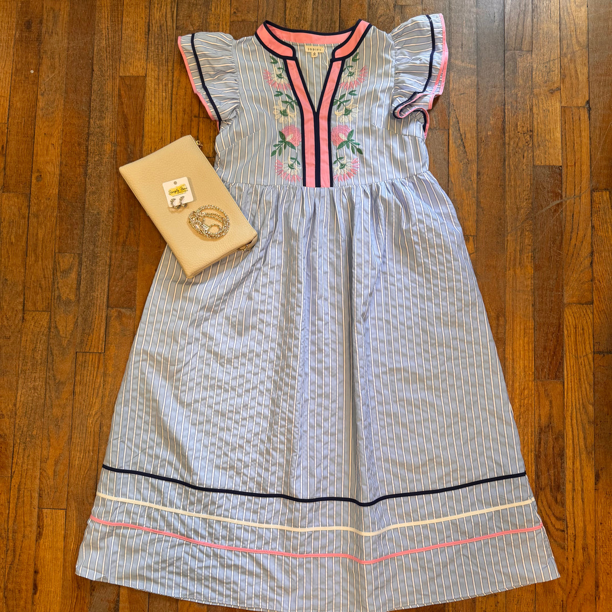 A light blue and white striped dress with pink and green floral embroidery on the chest, styled flat on a wooden floor next to a beige clutch bag.