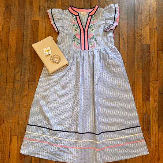 A light blue and white striped dress with pink and green floral embroidery on the chest, styled flat on a wooden floor next to a beige clutch bag.