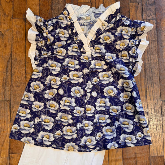 A navy blue floral top with white ruffle trim and a white V-neckline, laid flat on a wooden floor next to white pants.