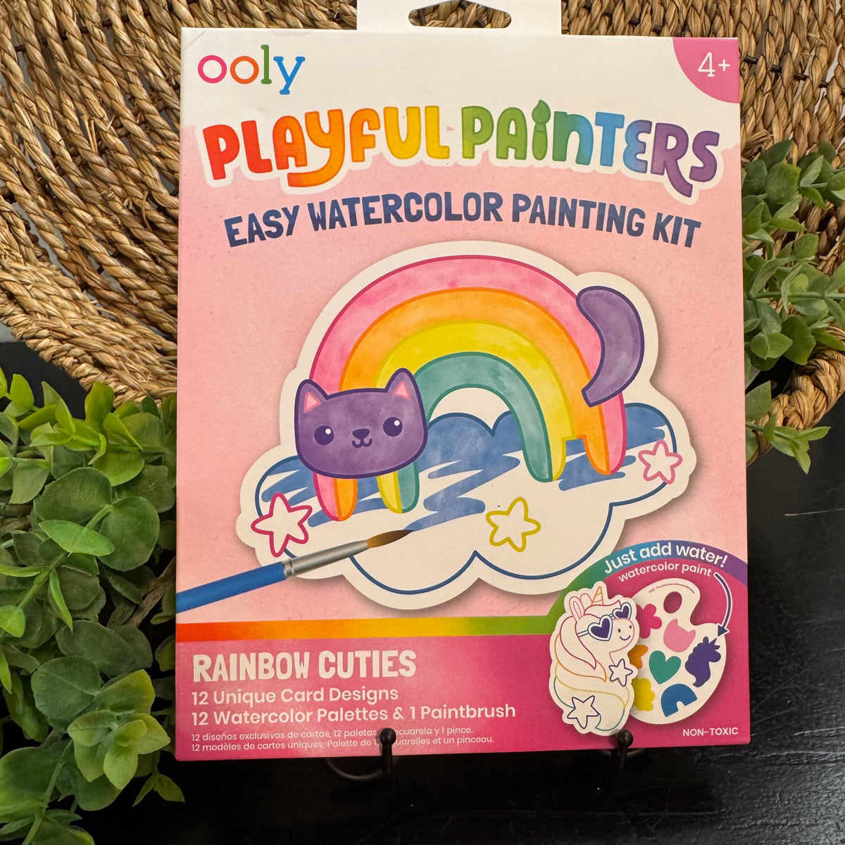 Ooly Playful Painters Easy Watercolor Painting Kit in its retail packaging, featuring a pink box with a colorful rainbow and purple cat illustration, displayed on a black stand with green plants in the background.