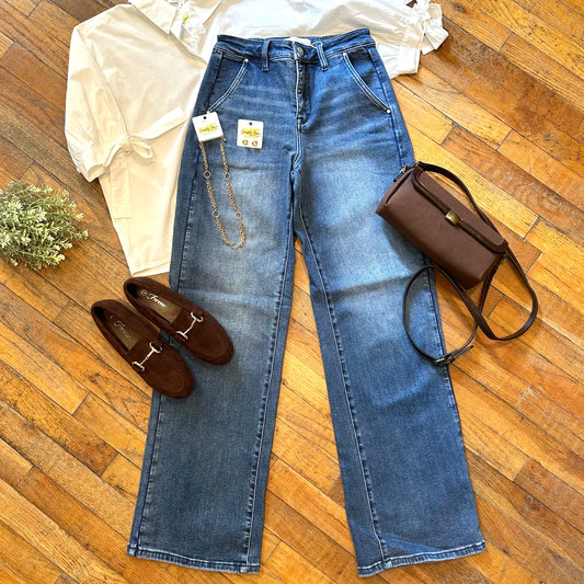 A pair of blue denim jeans laid flat on a wooden floor, styled with a white blouse, brown loafers, and a brown handbag. The jeans have a small gold chain detail attached near the waistband and are still showing their retail tags.
