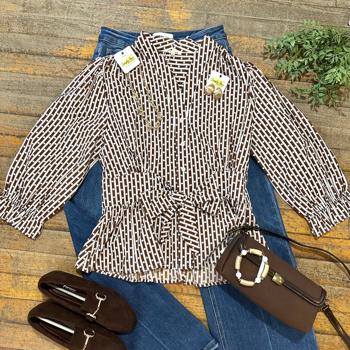 A women's long-sleeve belted blouse with a brown and white geometric pattern, styled in a flat lay on blue jeans with a brown handbag and brown loafers on a rustic wooden background.
